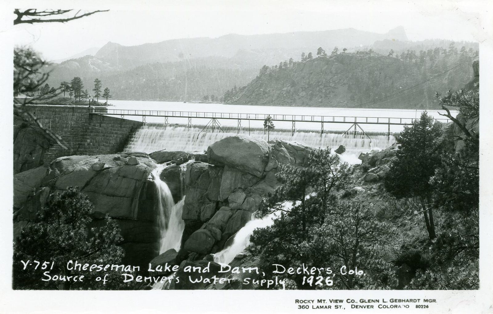 Water sheets over the full crest of a masonry dam into a granite gorge, a light service bridge spanning above it and the flat reservoir stretching back to timbered hills.