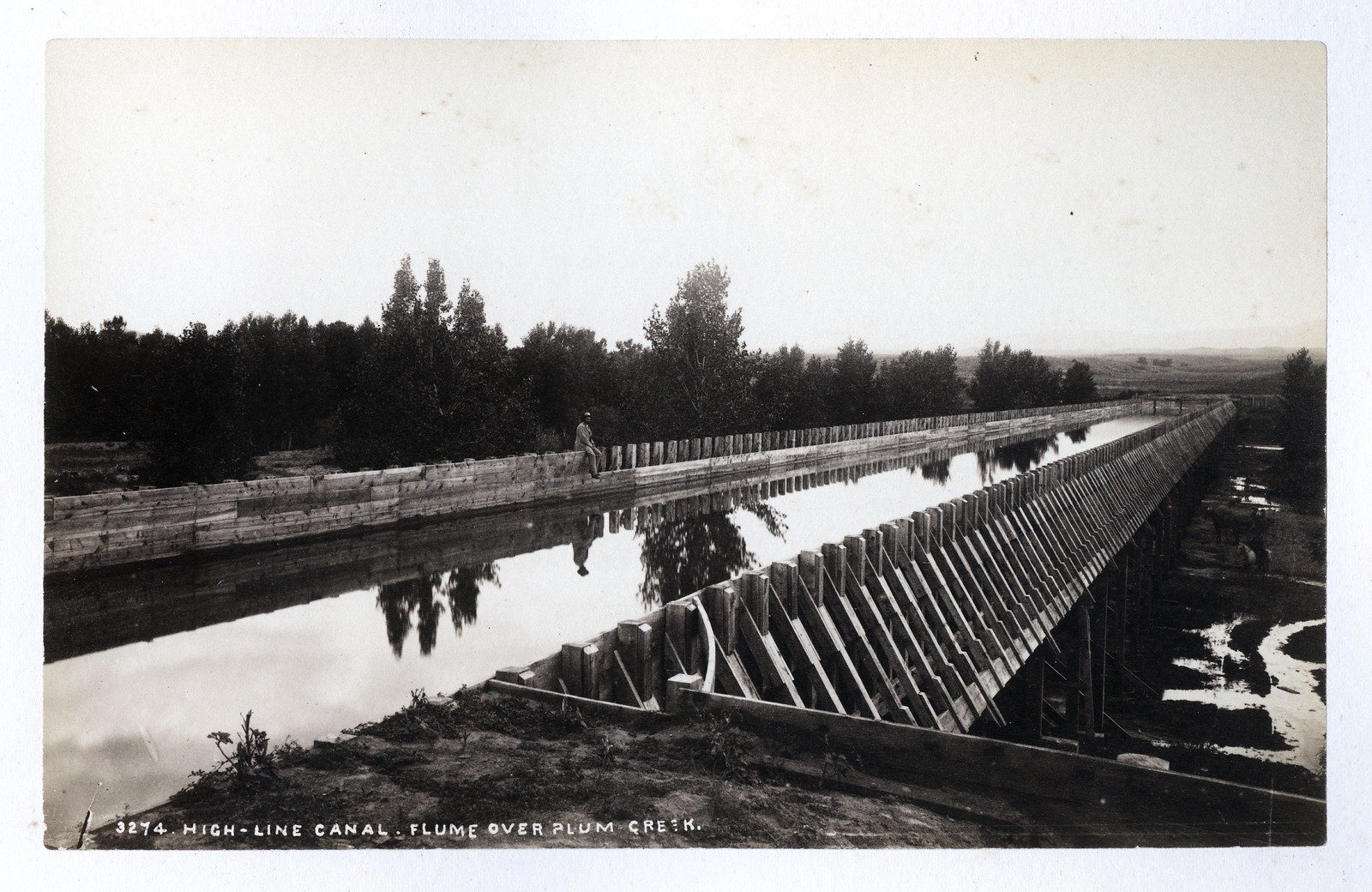 A long timber flume carries a canal on trestles across a creek bottom, its still water reflecting the trees on the bank, with a man sitting on the plank edge near the middle.