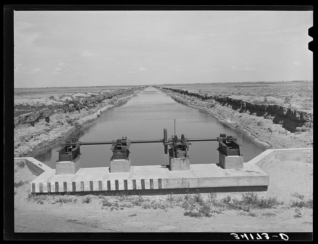 A straight canal full of water runs to the horizon across flat, bare farmland; in the foreground a concrete check structure carries four geared gate hoists on a single shaft.