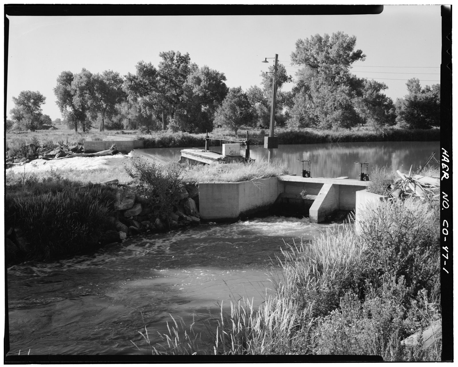 A low concrete headgate with two hand-wheel gate stands, water spilling through it into a grassy channel; behind, a river pooled above a diversion dam and a line of cottonwoods.