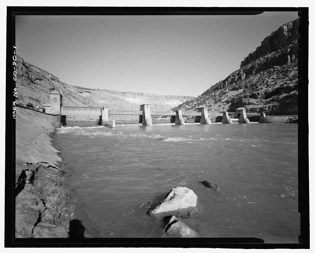 A long, low roller dam spans a wide river between canyon walls, its gate piers topped with small square towers; the river runs fast and pale over rocks in the foreground.