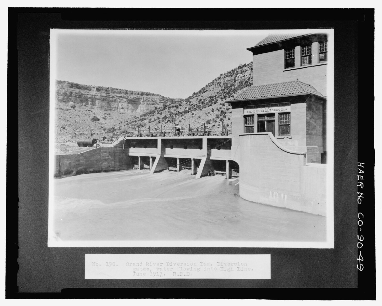 A print mounted on black card: water pours through the diversion gates beneath a concrete bridge deck into a canal, beside a tile-roofed gatehouse lettered GRAND RIVER DIVERSION DAM, with canyon cliffs behind.