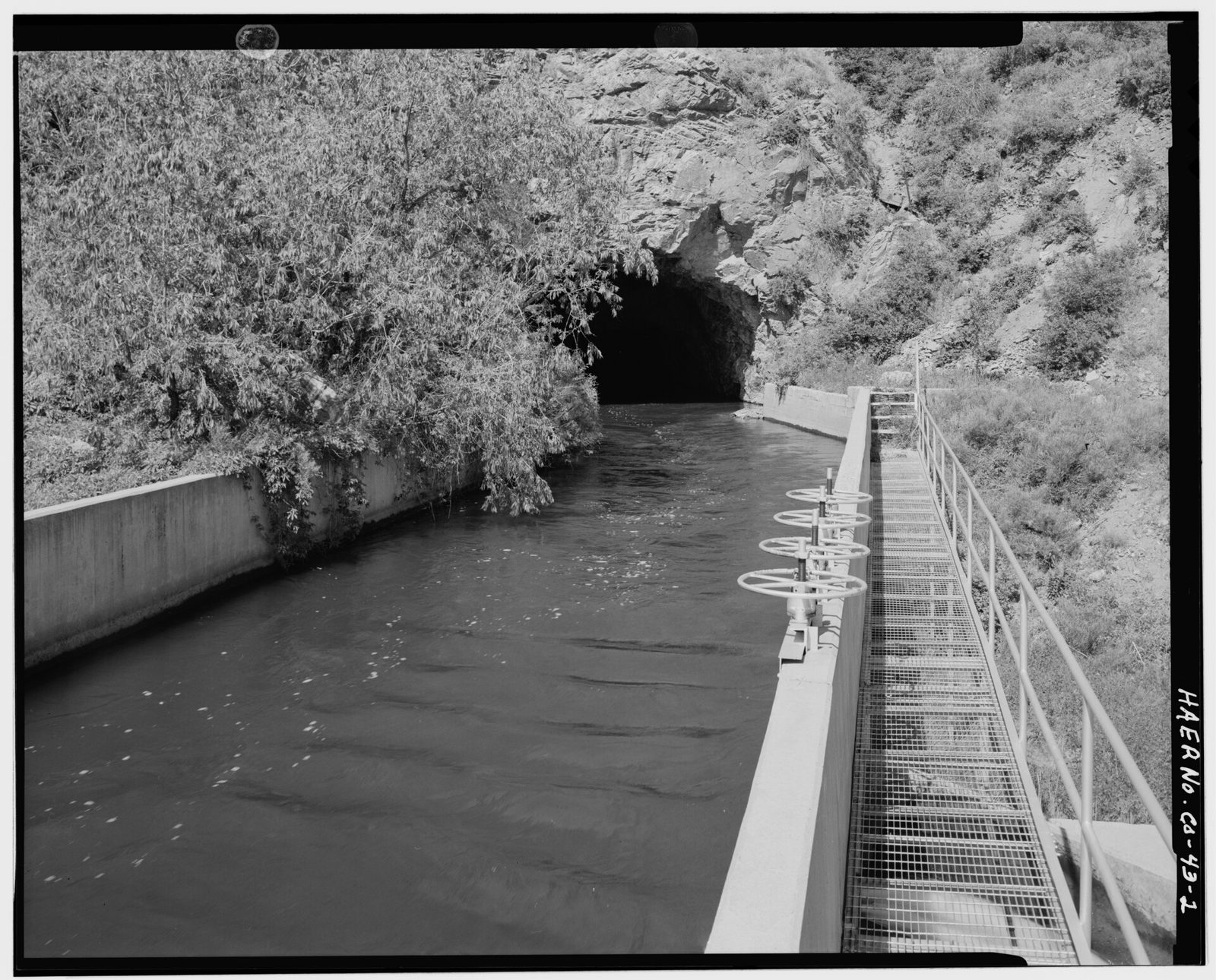 Canal exiting tunnel (left background) in Platte river canyon, with lateral gates in right foreground. View to west.