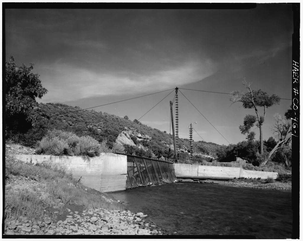 A low concrete diversion dam with a timber flashboard section crosses a river below a brushy hillside; a cableway tower and cables span the channel overhead, and cottonwoods stand on the far bank.
