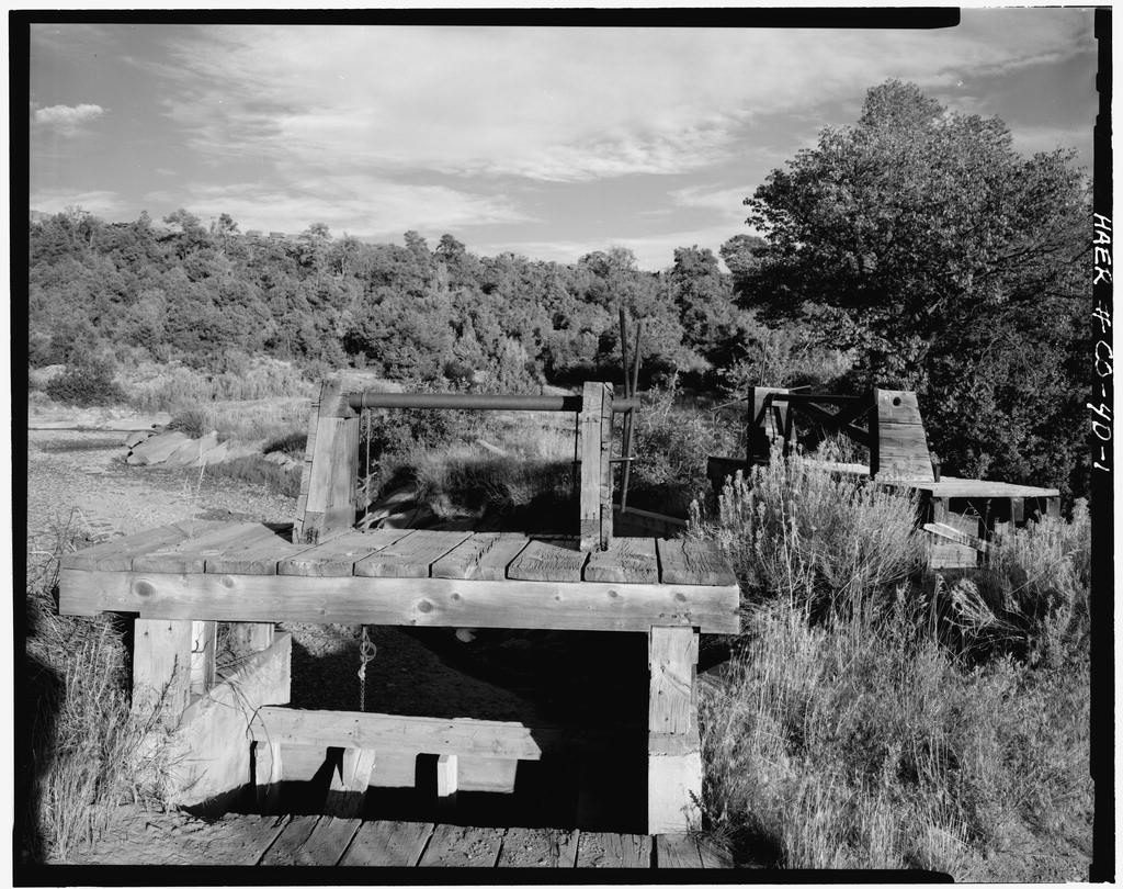 A weathered timber gate frame with a hand-crank shaft sits on a plank deck over a dry channel, sagebrush all round and a wooded ridge behind under a streaked sky.