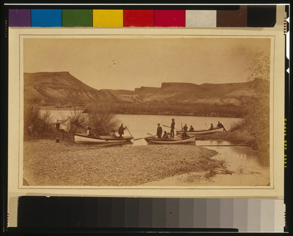 Three wooden boats drawn up on a gravel bar with men standing in them, flat-topped bluffs across the water behind, and a camera on a tripod under a dark cloth at the left edge.