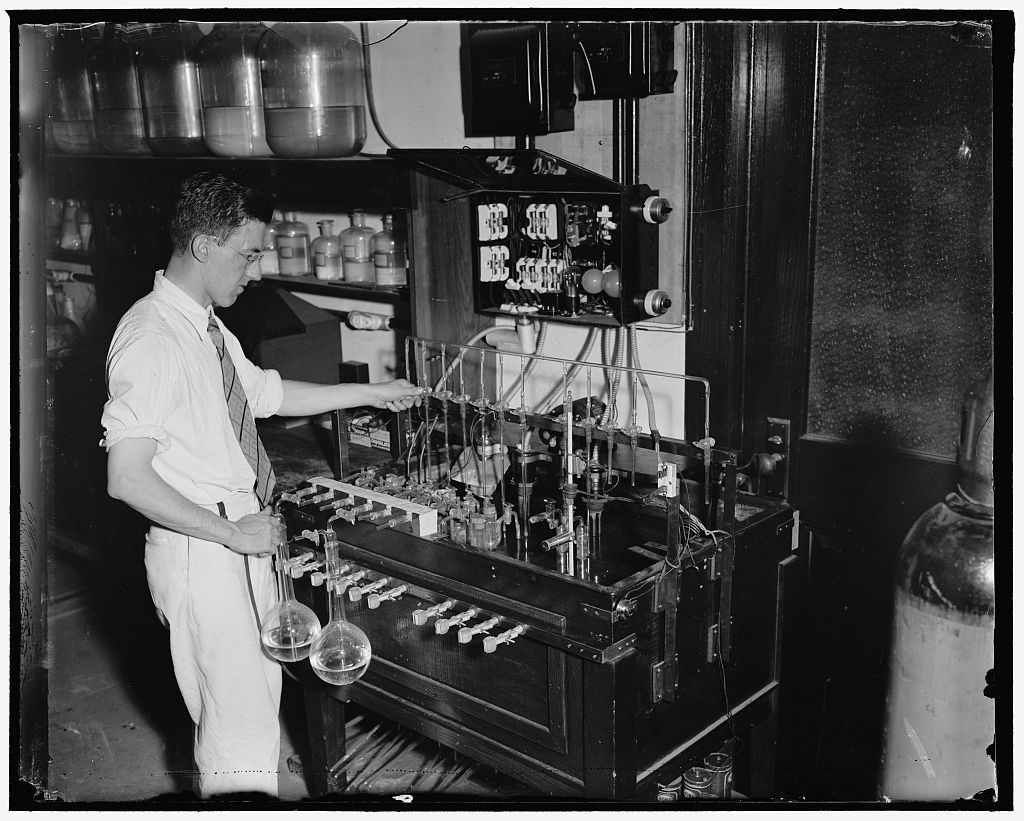 A chemist in shirtsleeves reaches across a bench of glass cells and rubber tubing, round-bottomed flasks hanging at its front and glass carboys ranked on the shelf above.
