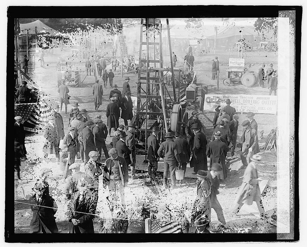 A crowd in hats and overcoats presses around a small drilling derrick on open ground at a show, a banner behind it advertising a well-drilling outfit.