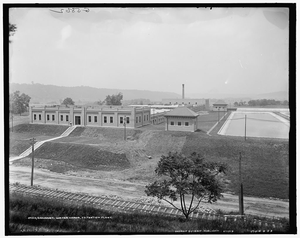 Long low brick filter houses stand on raw graded ground beside open rectangular settling basins, a railway line running across the foreground and a chimney on the skyline.