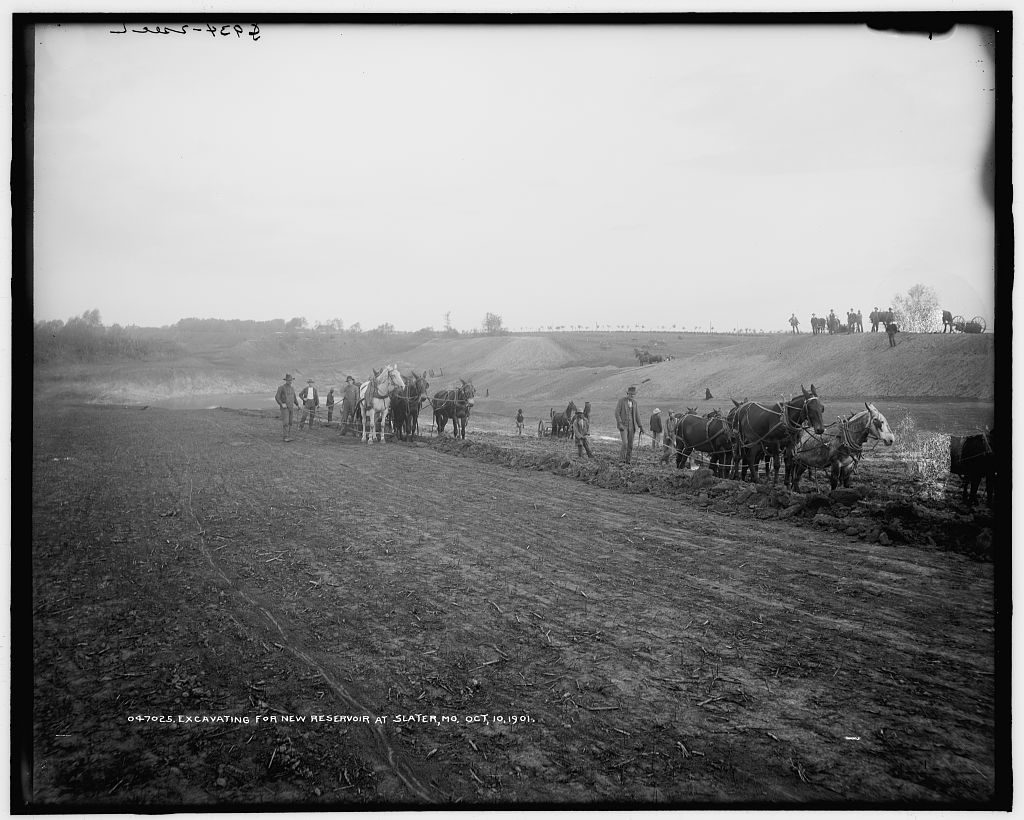 Horse teams and men stand on the scraped floor of a shallow excavation, spoil banked along one side and more teams working along the skyline.