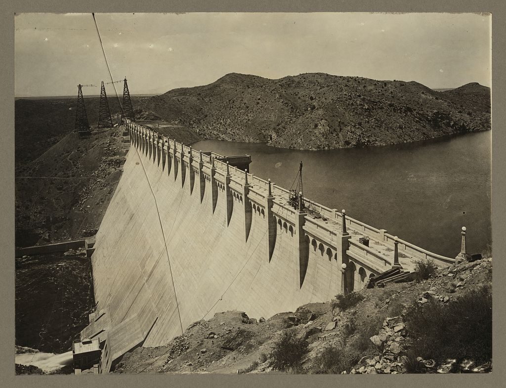 A long curved concrete dam seen down its crest, with a buttressed parapet and lamp standards, the reservoir filling behind it and cableway towers standing on the far abutment.
