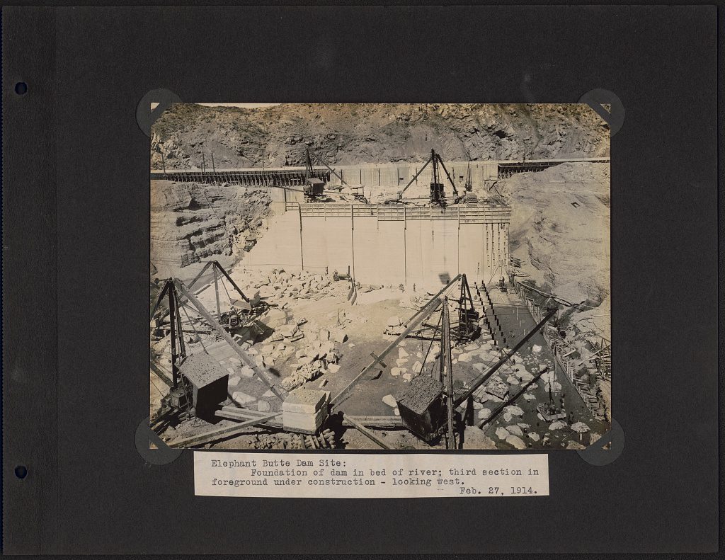 The dry bed of a river walled off for construction, with derricks and stiff-leg cranes standing over a poured foundation; the print is held to a black album page by corner mounts above a typed caption slip.