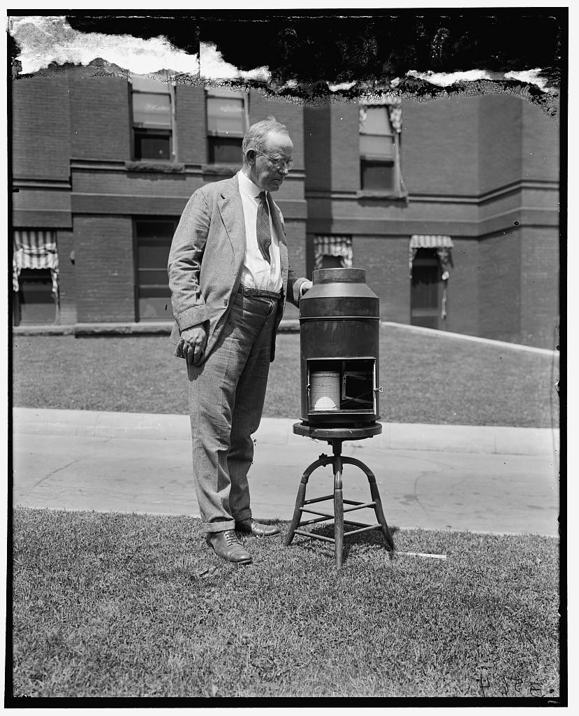 A grey-haired man in a suit stands on a lawn with one hand resting on a cylindrical recording gauge mounted on a cast-iron tripod, an open window in its side showing the chart drum.