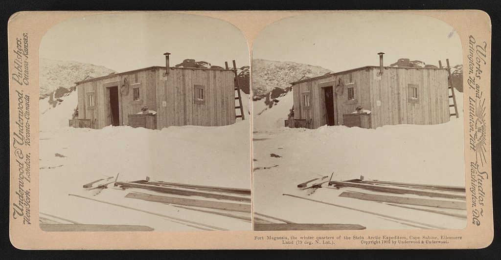 A stereograph card printing the same view twice side by side: a low plank hut half buried in a snowdrift, with sledge runners and lumber lying in the foreground.