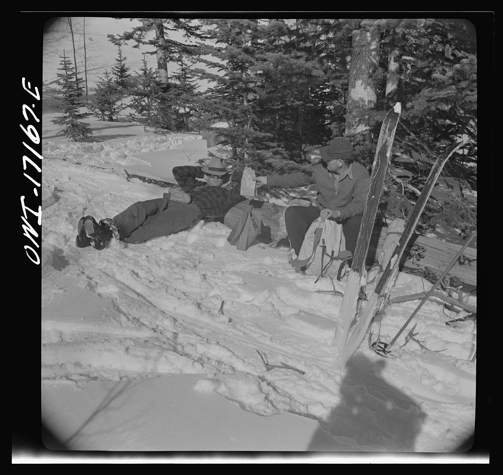 Two men rest on a packed snow trail in spruce timber, one lying back against his pack, with skis and poles standing upright in the snow beside them.
