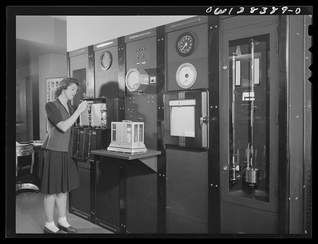 A woman marks a paper chart on a recording drum in a room lined with instrument cabinets, dial gauges, a clock and a glass-fronted case of barometric tubes.