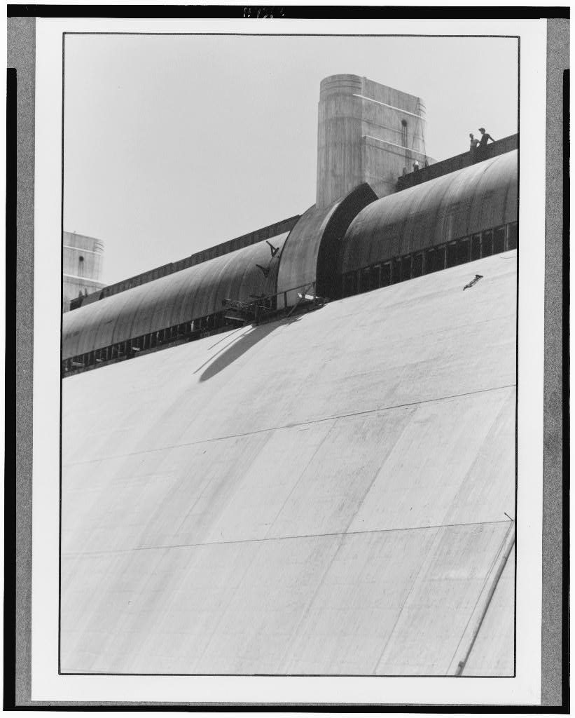 A vast curved steel drum gate lies along the lip of a blank concrete spillway apron, a square pier rising at its far end and one worker standing on the crest.