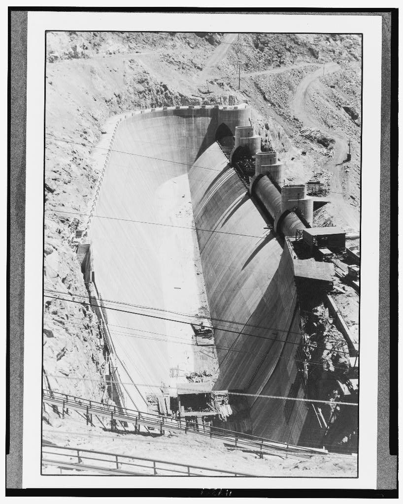 An enormous empty concrete spillway channel curves away between rock walls, drum-gate piers standing in a row along one side and figures tiny on the walkway at its foot.