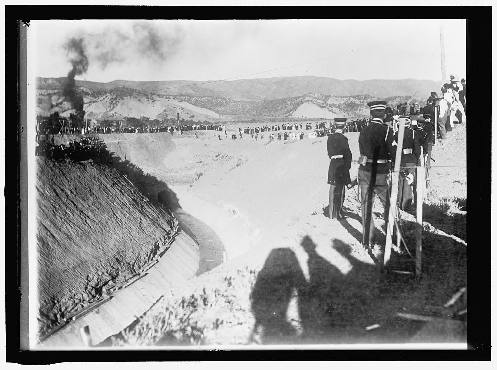 Uniformed officers stand in the foreground above a dry concrete-lined canal while a crowd lines its far rim, dry hills behind and a plume of black smoke rising at the left.