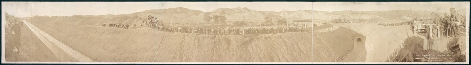 A long sepia panorama: a crowd lines the top of an earthen canal bank for hundreds of yards, a flag flies over a small grandstand, and the tunnel's arched portal opens at the right where the empty canal begins.