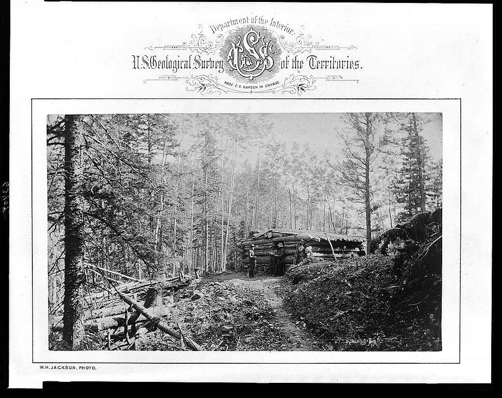 Three small figures stand at a low log cabin in dense standing timber, the print laid on a mount headed in ornate type 'U.S. Geological Survey of the Territories'.