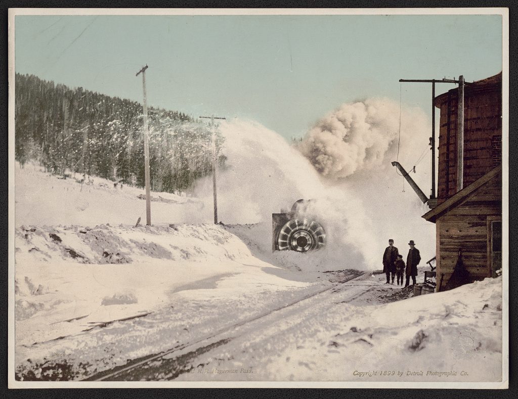 A rotary plough throws a high plume of snow across a cut track beside a timber water tower, with three onlookers standing clear and dark spruce on the slope behind.