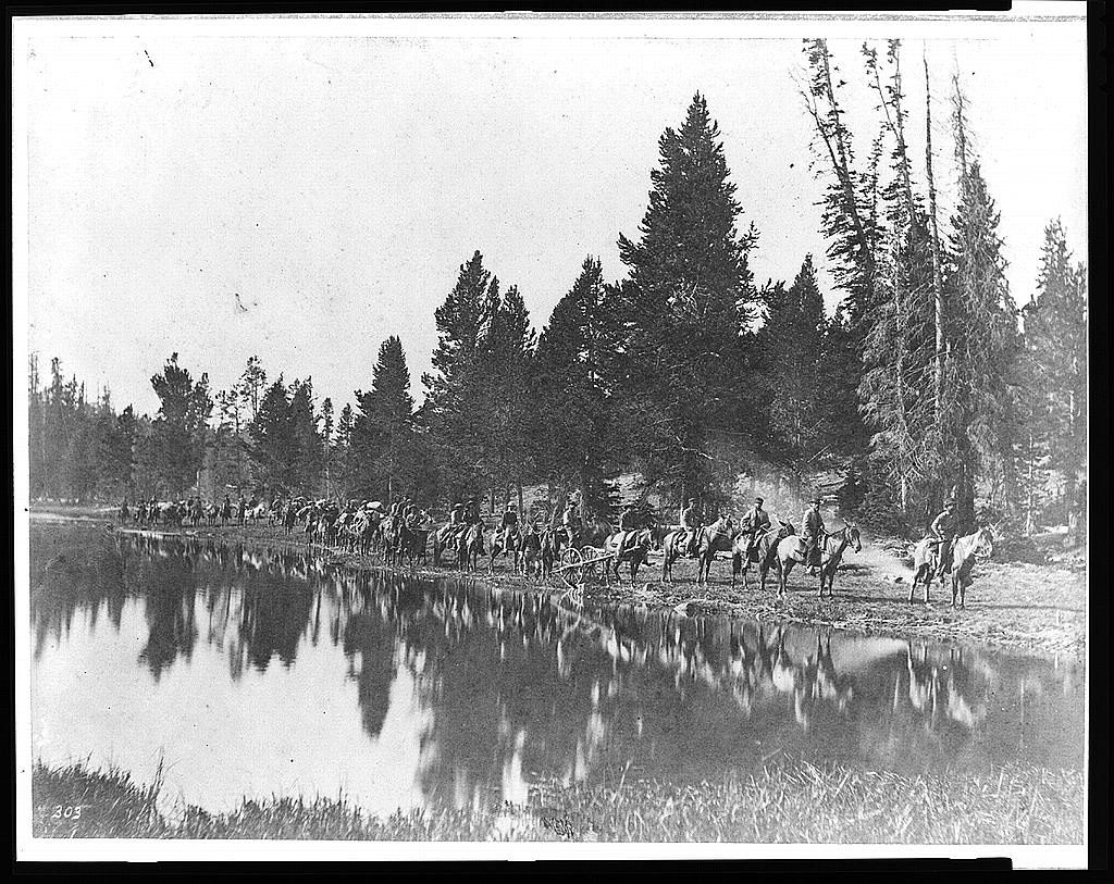 A long line of riders and pack animals strung out along a still riverbank, the whole train mirrored in the water, with spruce forest rising behind.