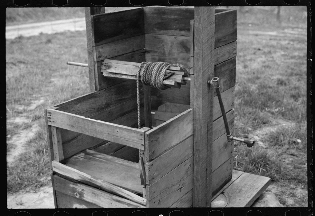 A rough plank well curb with a rope wound on a wooden windlass and an iron crank handle, standing open in grass beside a post.