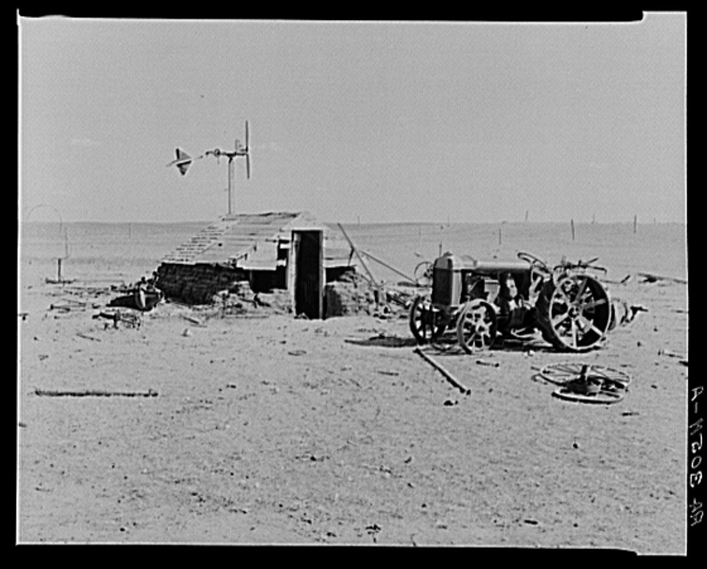 A tractor stands abandoned on bare drifted ground beside a collapsed machine shed, scattered ironwork around it and a windmill on the skyline.