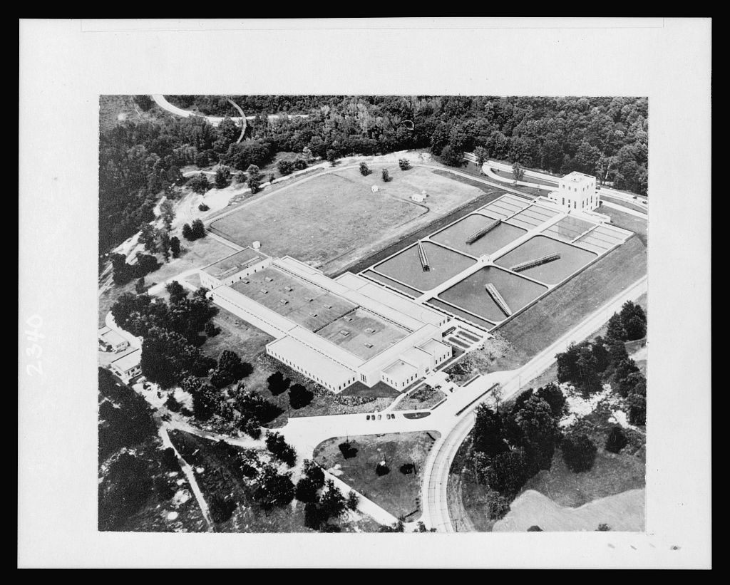 An aerial view of rectangular filter beds and long low plant buildings laid out in a clearing among woods, with a curving road along one edge.