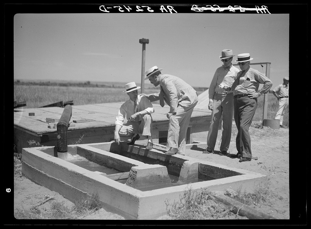 Four men in straw hats and shirtsleeves stand around a concrete stilling basin on flat open plains while water pours into it from a pipe.