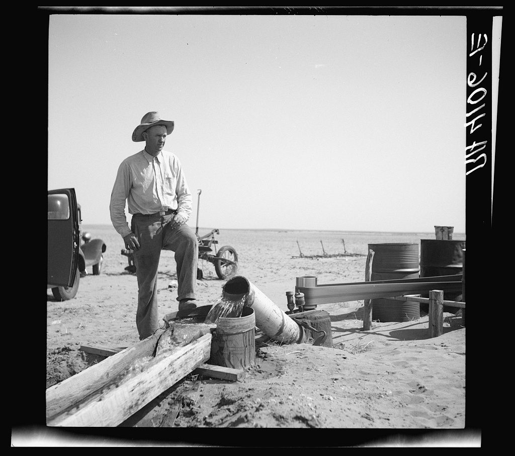 A farmer in a wide hat stands over a discharge pipe pouring water into a plank flume on bare blown sand, with oil drums, a car and a flat empty horizon behind.