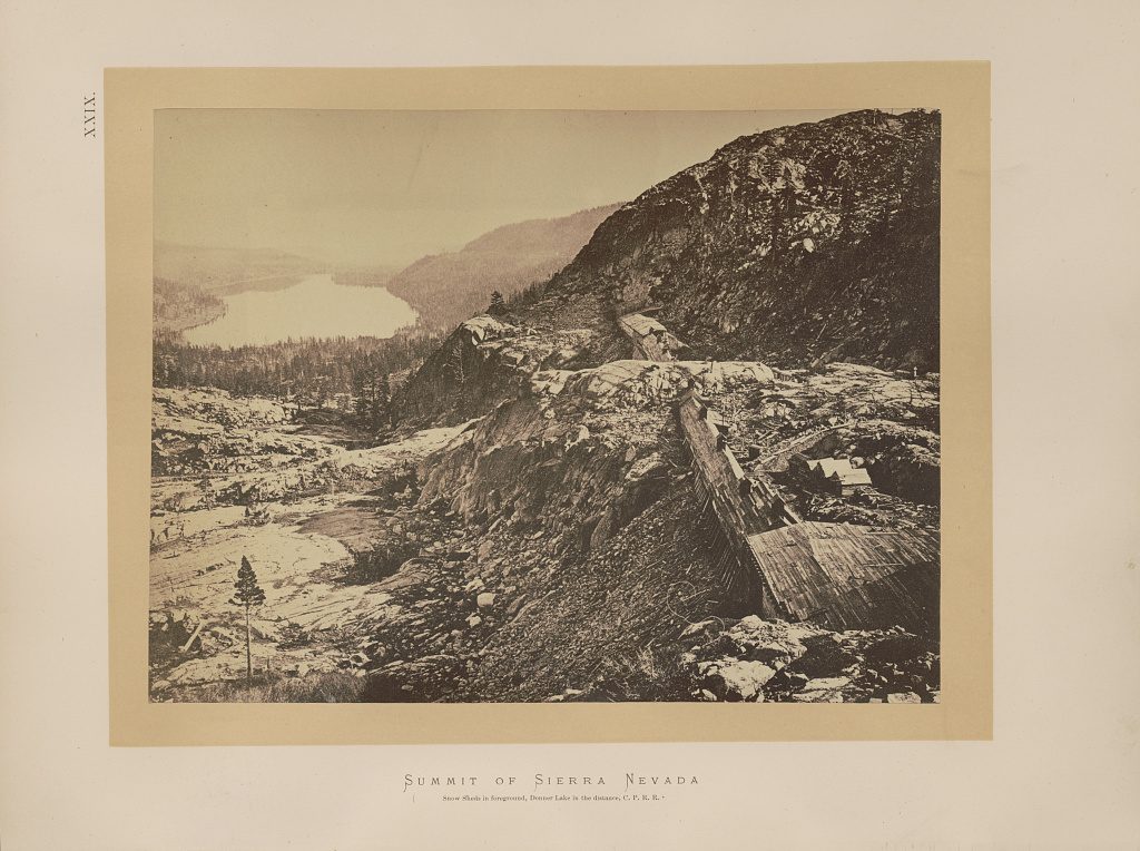 Timber snowsheds run down a bare granite slope between tunnel mouths, with a long lake in the valley far below and no snow anywhere in the frame.