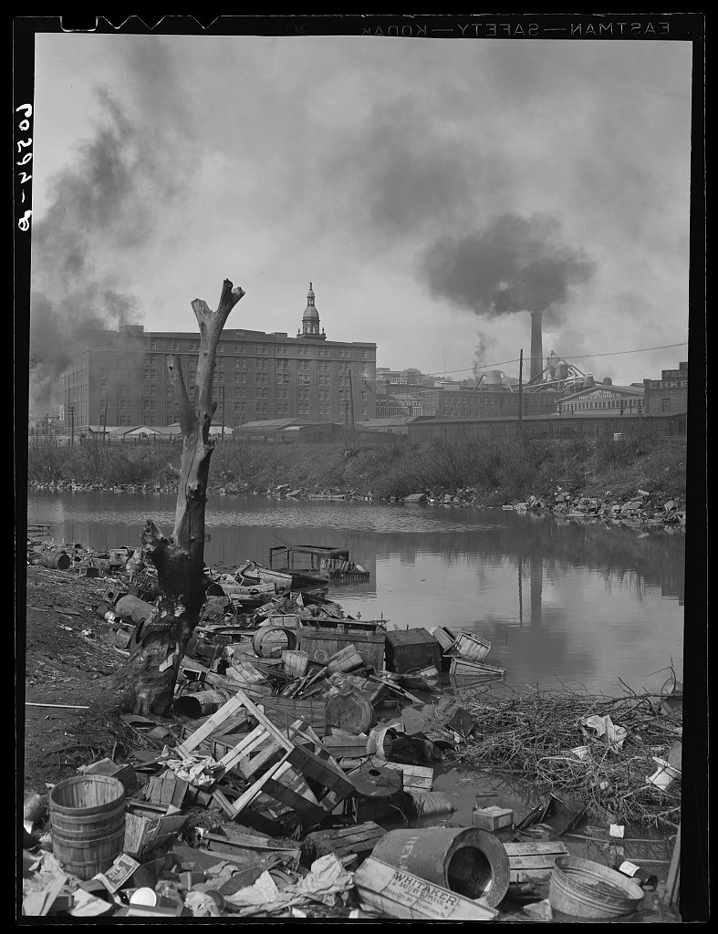 A still river backwater with its bank heaped in tins, crates, barrels and a washtub, a dead stump standing in the foreground and factory chimneys pouring black smoke behind.