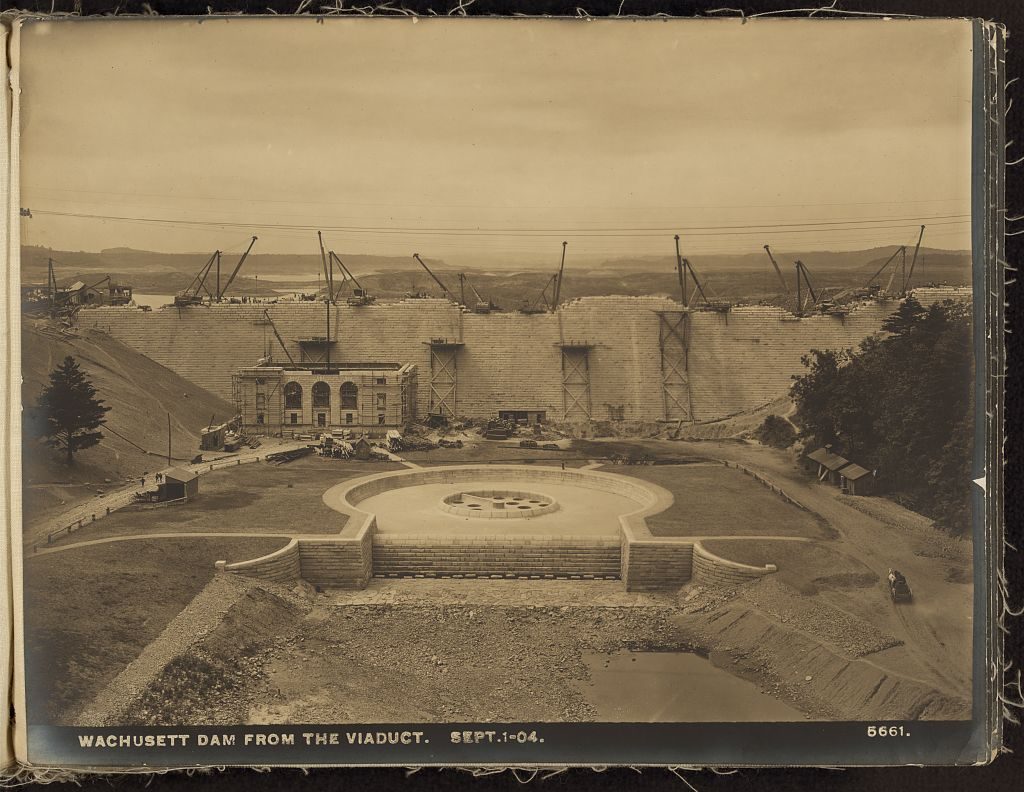 A masonry dam half built across a valley, derricks standing along its crest, with a finished stone gatehouse and a formal landscaped basin laid out below it.