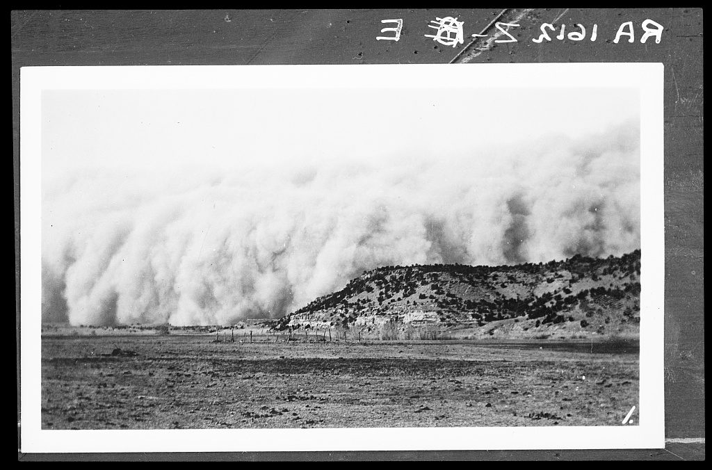 A towering wall of dust rolls across the plains and over a low juniper-topped mesa, with bare scoured ground in the foreground.
