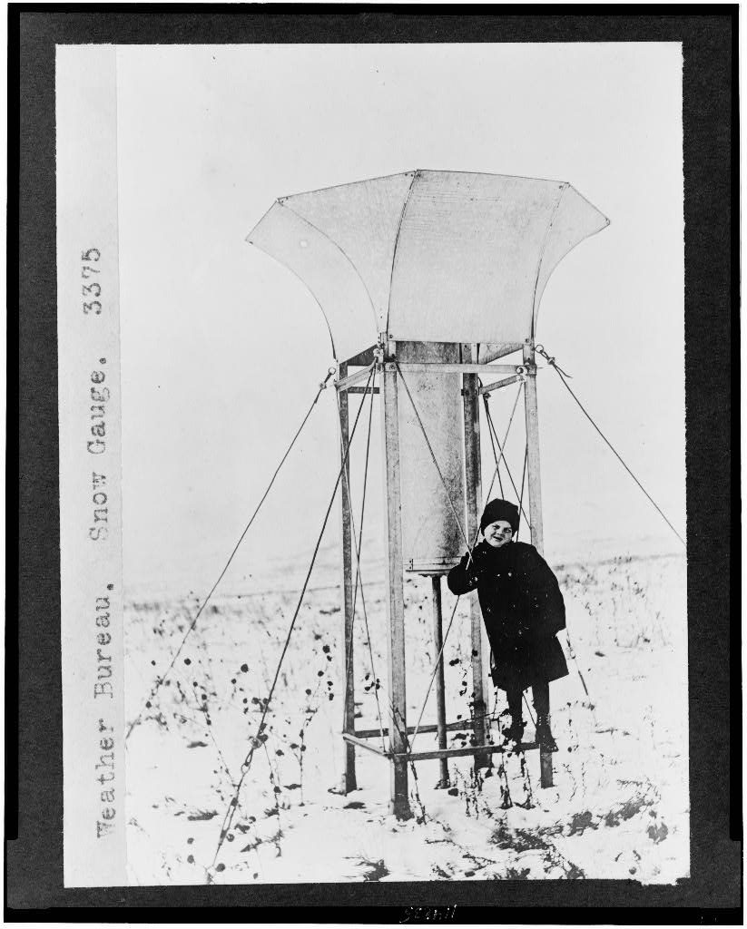 A child in a heavy coat and cap stands on the frame of a tall funnel-mouthed gauge, guyed with wires, alone in a snow-covered field.