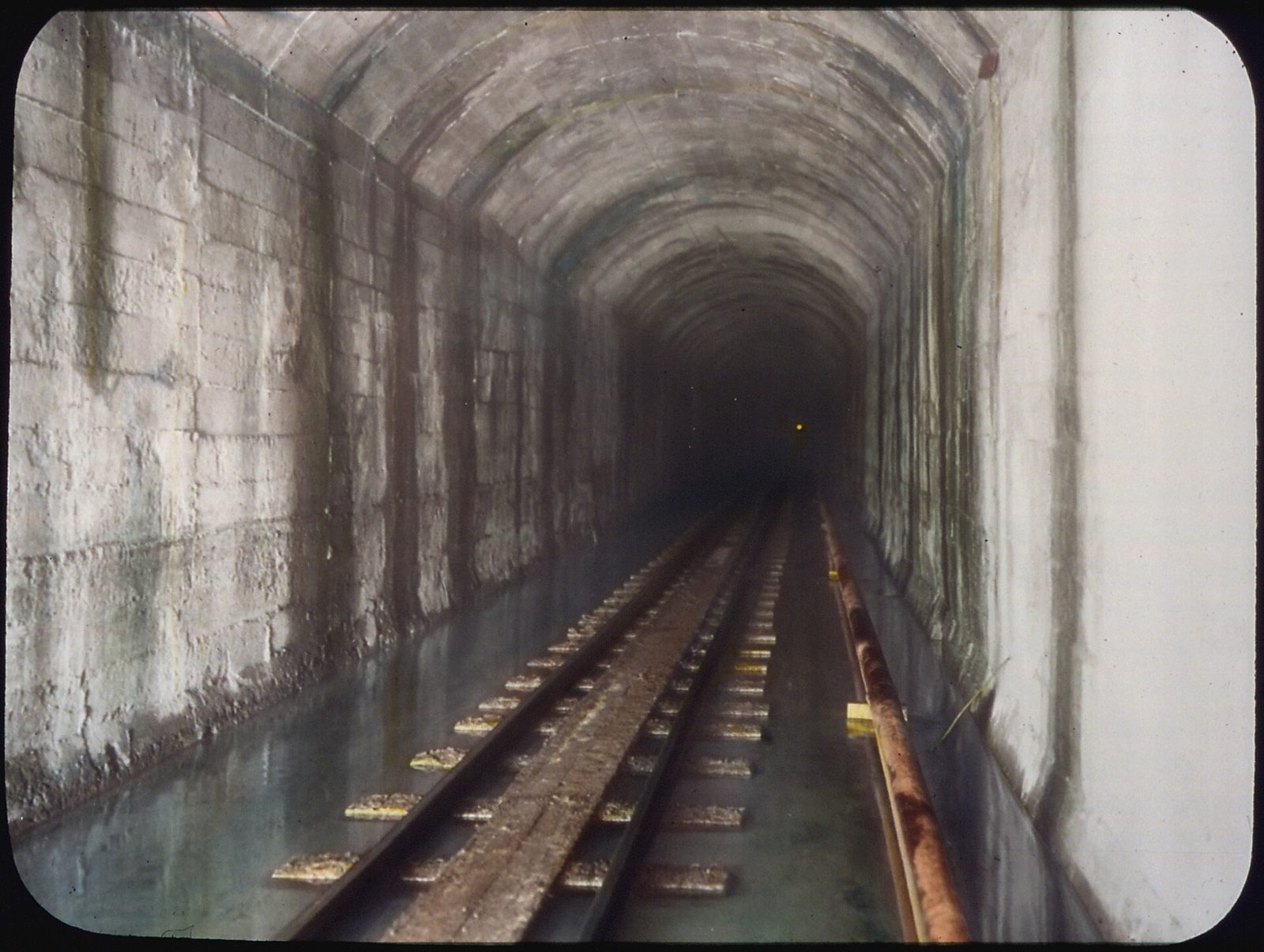 A hand-coloured lantern slide looking down a finished section of concrete-lined tunnel; a narrow-gauge construction track runs along the floor into darkness toward a single distant light.