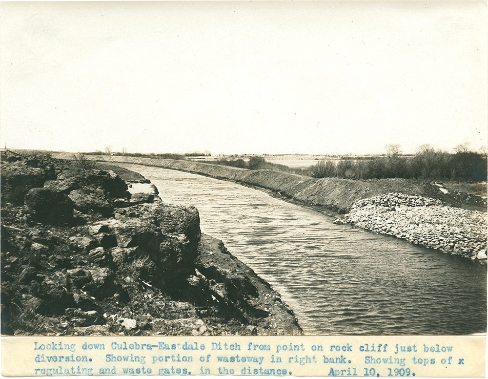 A sepia print of a wide new canal curving away across flat country, seen from a rocky bluff; a rock-armoured wasteway sits on the far bank. A typed caption is pasted below the image.