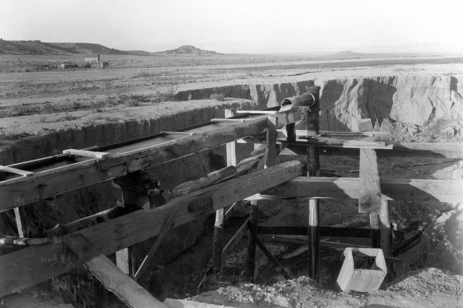 A discharge pipe runs out over a timber-framed trough at the head of a raw eroded gully, with a small square measuring box set on the ground beside it and open plain behind.