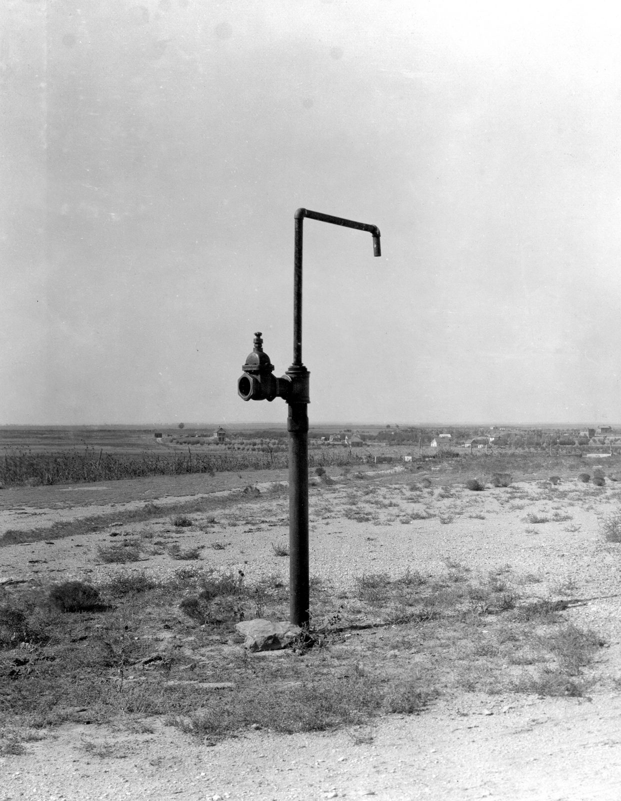 A single capped standpipe with a gate valve stands alone on bare short-grass plain, a right-angled spout at the top, with flat farmland and a far horizon behind.