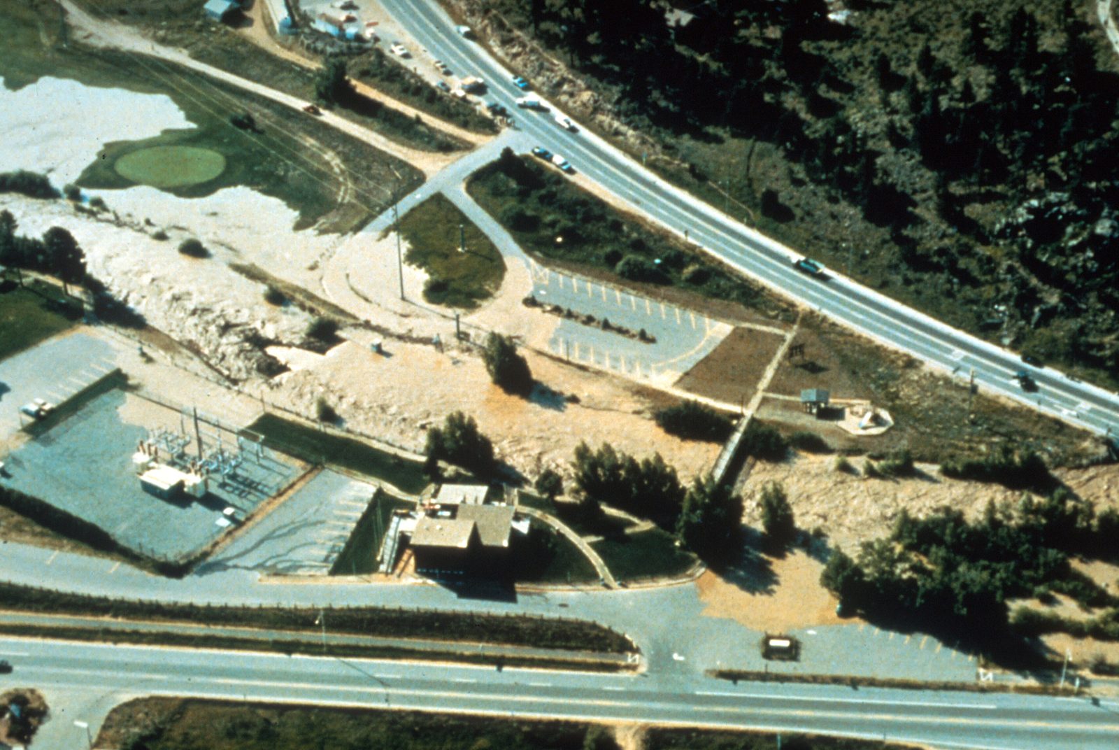 An aerial view of a muddy river running through a town, pale sediment spread in fans across a golf course and roads, with buildings and traffic alongside.