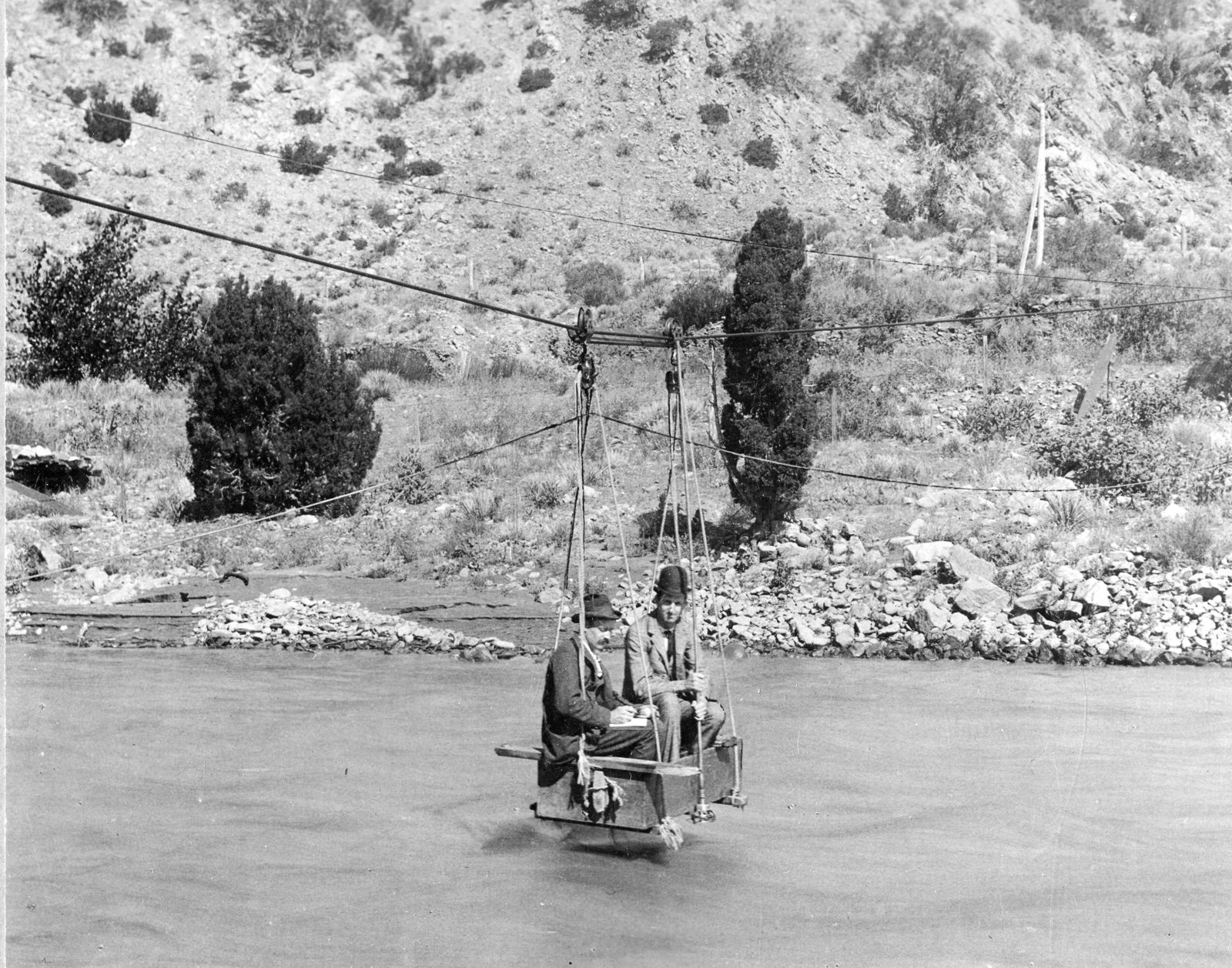 Two men in hats sit in a small open car slung from a cable above a wide river, one working a reel over the side, with a scrub-covered canyon slope behind.