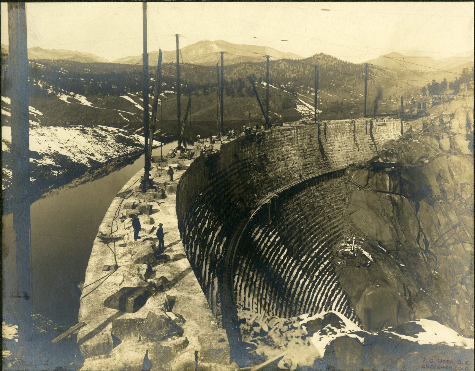 A curved masonry dam rising from bare granite, its stepped inner face half built, with derrick masts standing along the crest, workmen on the stonework and forested hills behind.