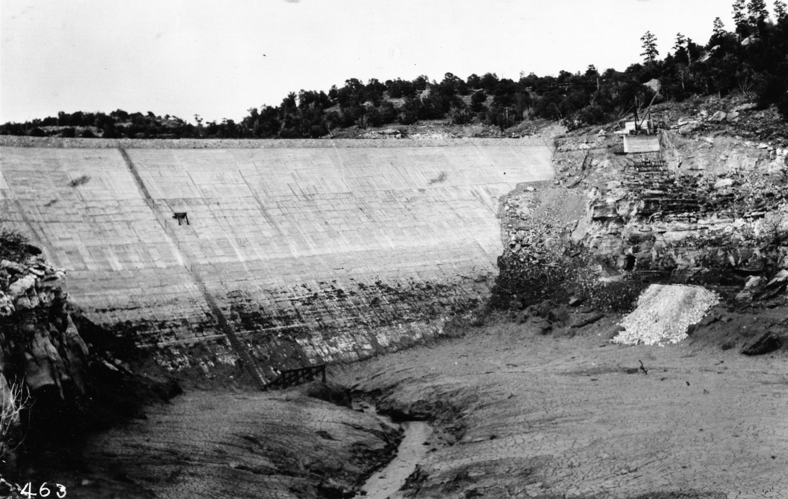 A dam with a smooth paved upstream face rising from an empty basin, a rock outcrop and an outlet structure at its right end, and piñon on the slopes above.