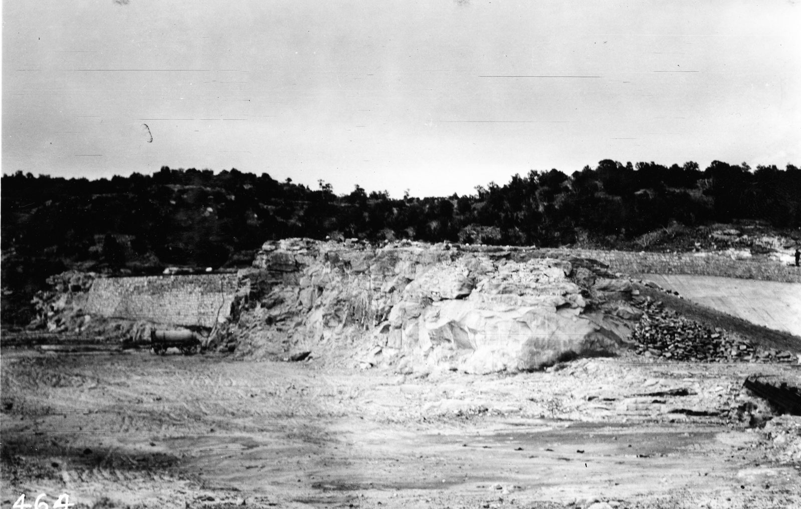 A broad spillway channel excavated through pale rock, a low embankment closing the ground at the right, and the dry floor of the reservoir basin in the foreground.