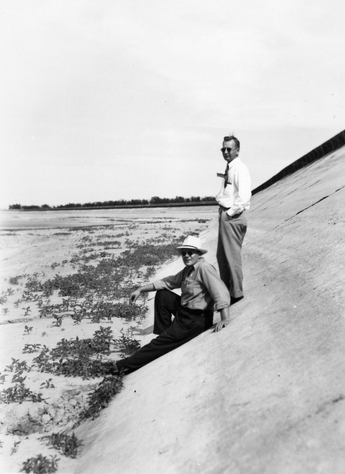 Two men in shirtsleeves stand and sit on the smooth sloping concrete face of a reservoir lining, weeds growing out of the cracked flat bed stretching away behind them.