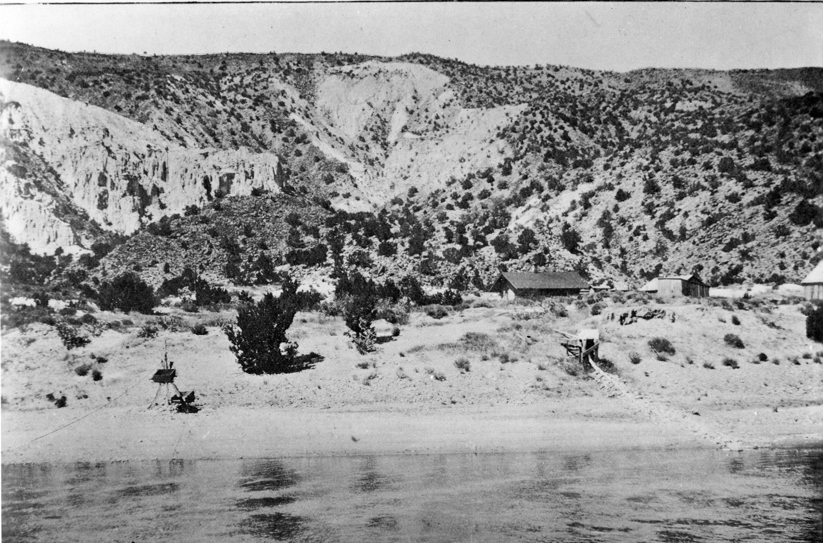 A shallow river runs past a pale sand bar below juniper-dotted bluffs, with two small pitched-roof huts and a cable rig standing on the bank above the water.