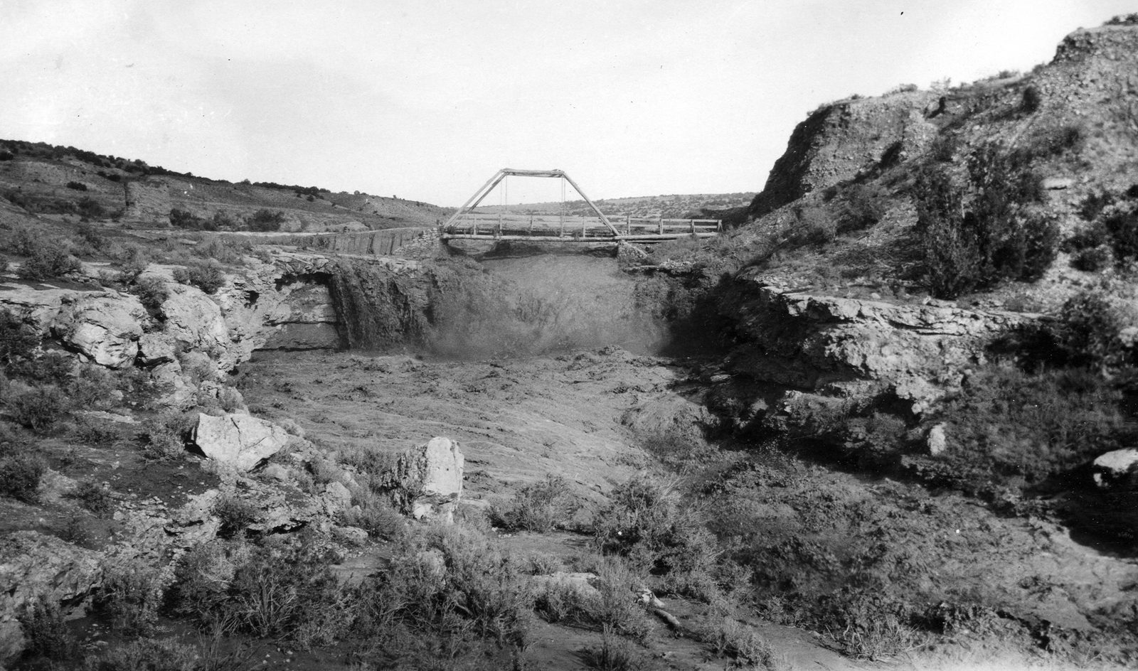 Muddy water pours through a narrow slot cut in pale sandstone beneath a light timber truss bridge, dry sage benches rising on both sides.