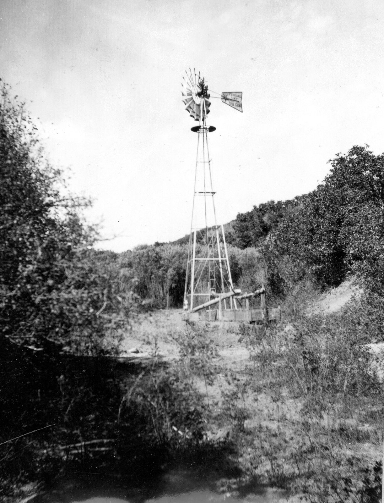 A tall lattice windmill tower rising above brush in a narrow wooded canyon, a wooden trough and platform at its foot and trees close on either side.